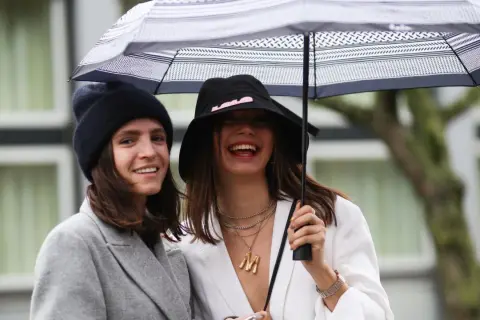 Dos mujeres ríen bajo un paraguas, luciendo un estiloso outfit lluvia mujer con gorros y abrigos.