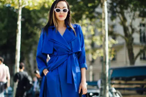Mujer con gabardina azul eléctrico, gafas blancas y bolso negro, paseando por la calle.