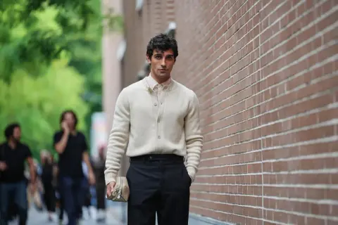 Hombre con suéter crema y pantalón negro, bolso de mano beige. Un ejemplo de las últimas tendencias moda hombre.