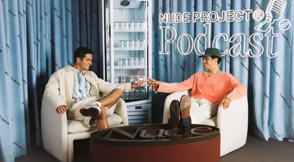 Los fundadores de Nude Project brindan con latas de su bebida, sentados en sillones frente a un cartel de neón del podcast de la marca.