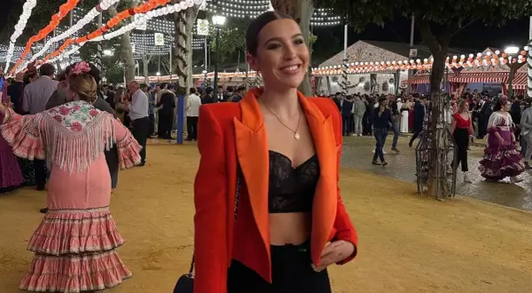 Chica sonriente con chaqueta naranja y top negro, lista para la ropa para ir a la feria de dia. Ambiente festivo con luces y gente.