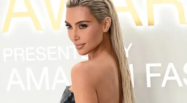 Kim Kardashian con pelo efecto mojado, luciendo un vestido negro brillante. El fondo tiene letras doradas y blancas.