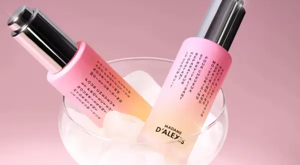 Dos frascos de aceite para pelo, con un degradado rosa y amarillo, reposan sobre cubitos de hielo en una copa.