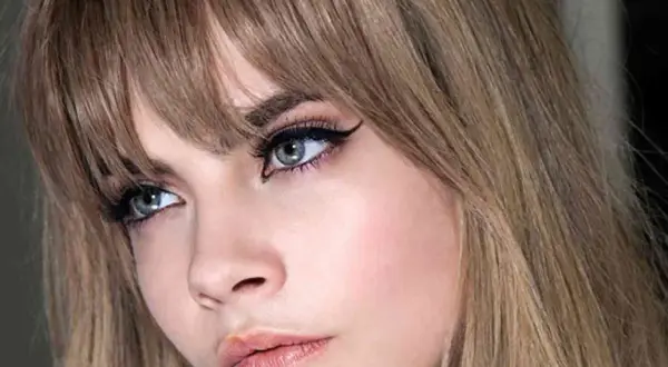 Primer plano de una mujer con flequillo rubio desfilado, delineado de ojos dramático y labios rosados.