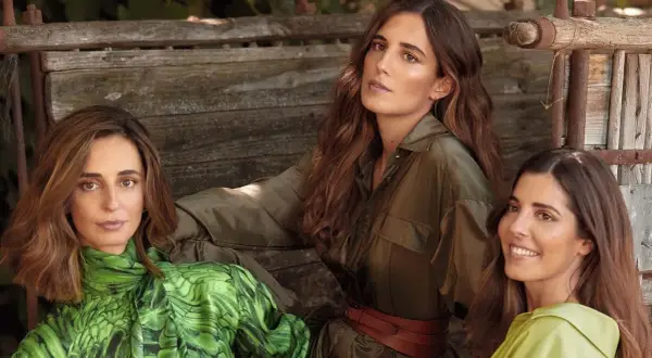 Belen Corsini y dos mujeres más, vestidas con ropa de tonos verdes, posan sentadas en un banco de madera rústico.