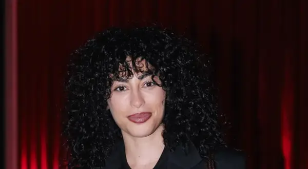 Mina El Hammani, con su cabello rizado y oscuro, sonríe ante un fondo de cortinas rojas.