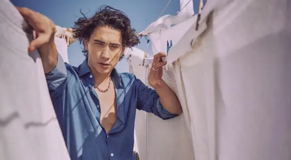 Enzo Vogrincic, con su camisa desabrochada y collar llamativo, posa entre sábanas blancas tendidas al sol.