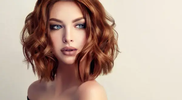 Mujer con cabello cobrizo ondulado en un elegante corte midi. Sus ojos azules y labios definidos resaltan su belleza.