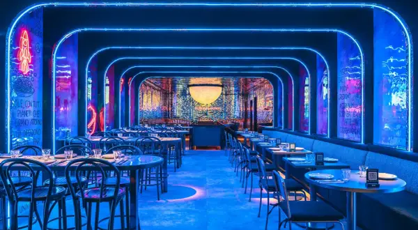 Un restaurante futurista con luces de neón azules y arte mural. Mesas puestas para cenar, quizás para disfrutar de una beata pasta bajo las estrellas.