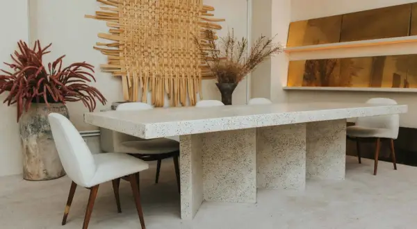 Comedor con mesa de terrazo y sillas tapizadas, arte de pared tejido y jarrones con flores secas. Un ambiente de lujo silencioso.