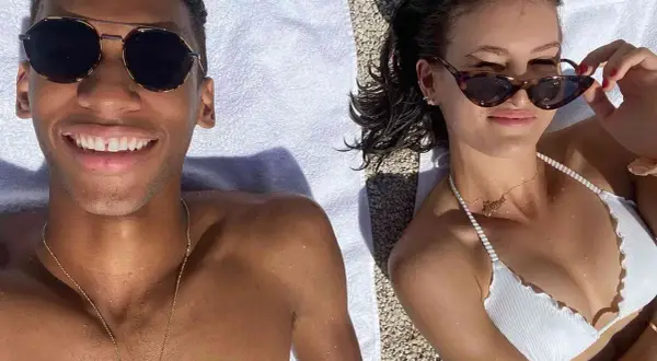 Felix Auger-Aliassime y su novia disfrutan del sol en la playa. Él sonríe a la cámara, ella se ajusta las gafas de sol.