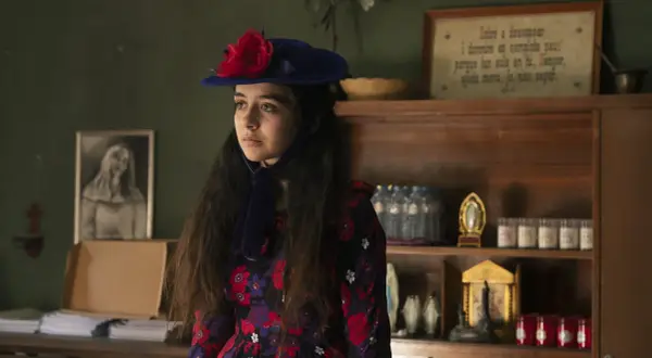 Irene Balmes, con un sombrero azul y un vestido floral, mira pensativa. Detrás, estanterías con objetos religiosos y un cuadro.