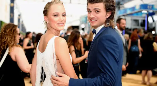 Joe Keery y su pareja, ambos sonriendo a la cámara en una alfombra roja. Ella lleva un vestido blanco con flecos, él un traje azul.