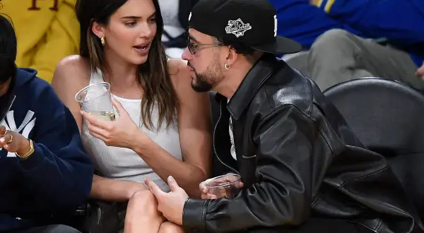 Bad Bunny y Kendall Jenner conversan en un evento deportivo, ella con un vaso en la mano y él con chaqueta de cuero.