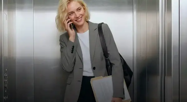 Mujer sonriente en ascensor, con un elegante outfit oficina, hablando por teléfono y cargando carpetas.