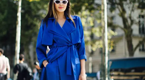 Mujer con gabardina azul eléctrico, gafas blancas y bolso negro, paseando por la calle.