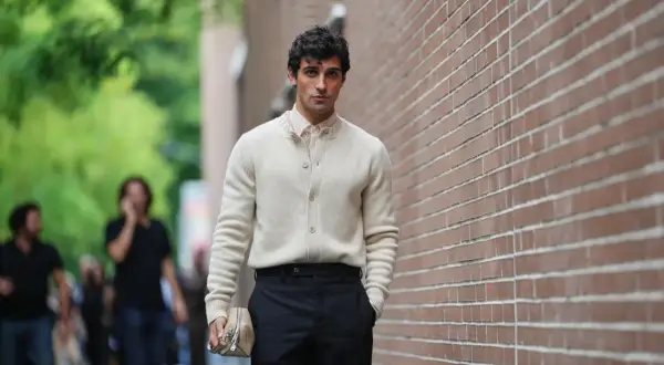 Hombre con suéter crema y pantalón negro, bolso de mano beige. Un ejemplo de las últimas tendencias moda hombre.