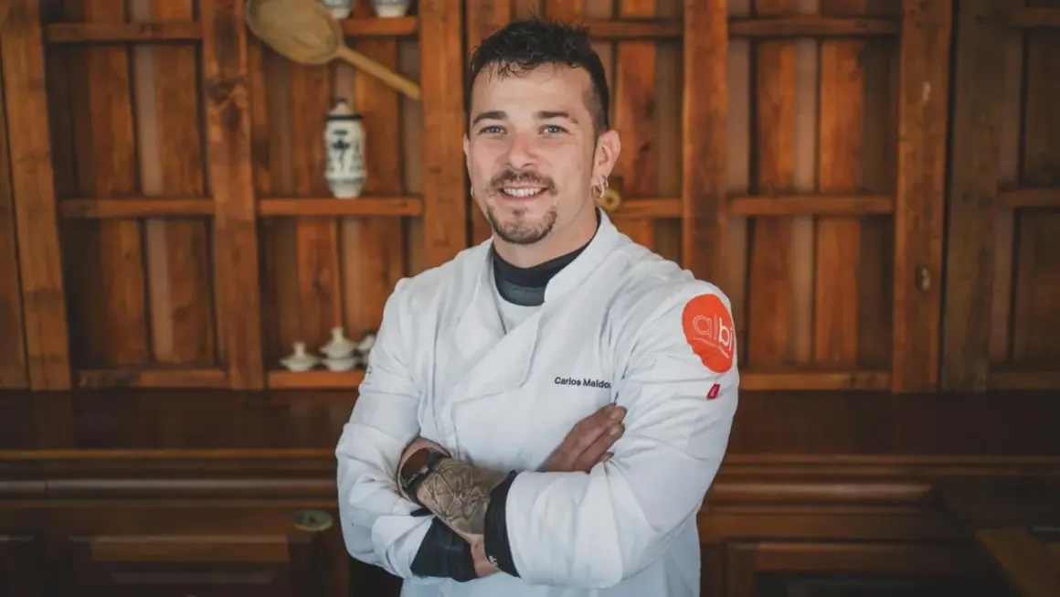 Carlos Maldonado, chef con tatuajes y barba, sonríe con los brazos cruzados frente a una estantería de madera.