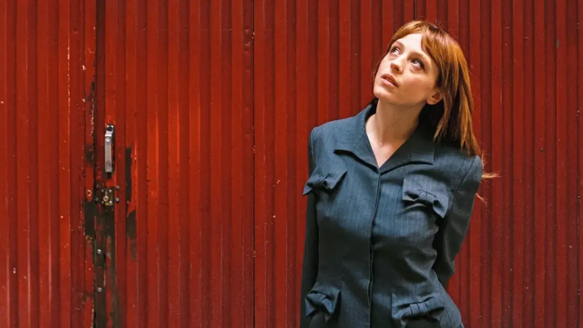 Catalina Sopelana, con su cabello rojizo y mirada perdida, posa frente a una puerta roja acanalada, luciendo un traje gris con detalles de lazos.