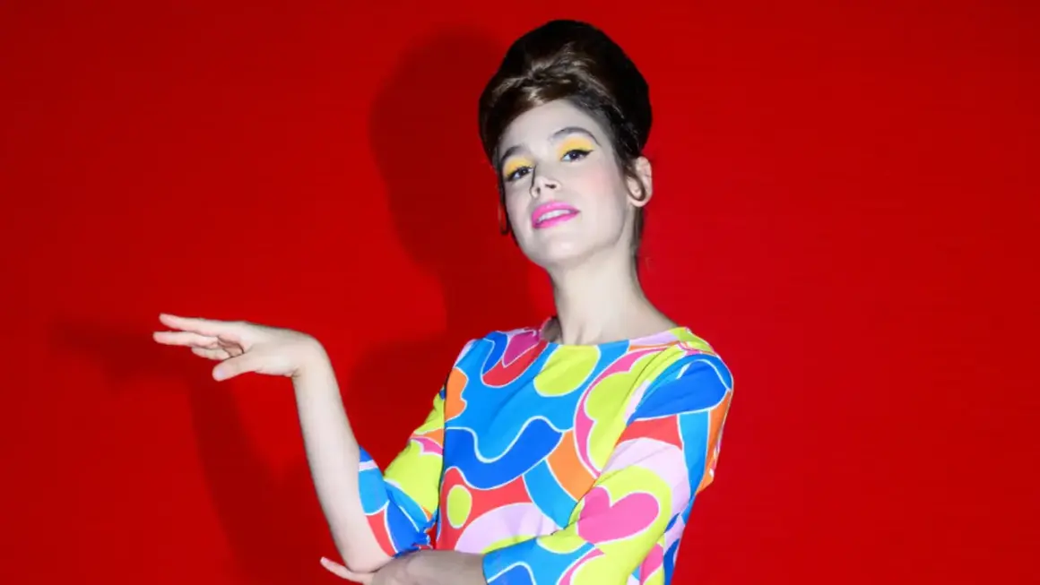 Cosima Ruiz de la Prada, con un peinado voluminoso y maquillaje vibrante, posa con un vestido de estampado psicodélico sobre fondo rojo.