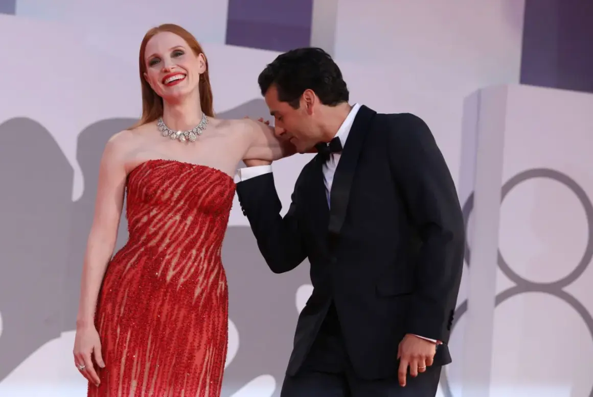 Oscar Isaac besa la mano de Jessica Chastain, quien luce un vestido rojo brillante y un collar de diamantes.