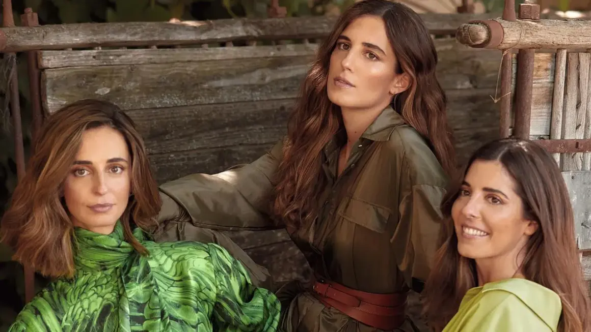 Belen Corsini y dos mujeres más, vestidas con ropa de tonos verdes, posan sentadas en un banco de madera rústico.
