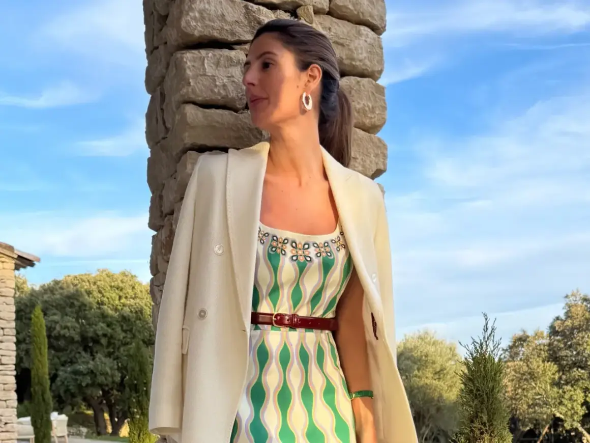 María de la Orden, con un vestido estampado y blazer blanco, posa junto a una pared de piedra bajo un cielo azul.