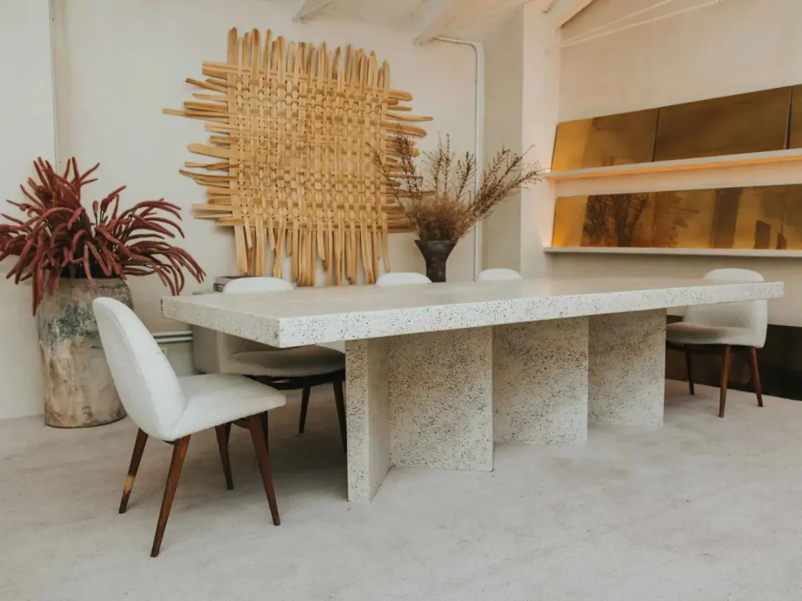Comedor con mesa de terrazo y sillas tapizadas, arte de pared tejido y jarrones con flores secas. Un ambiente de lujo silencioso.