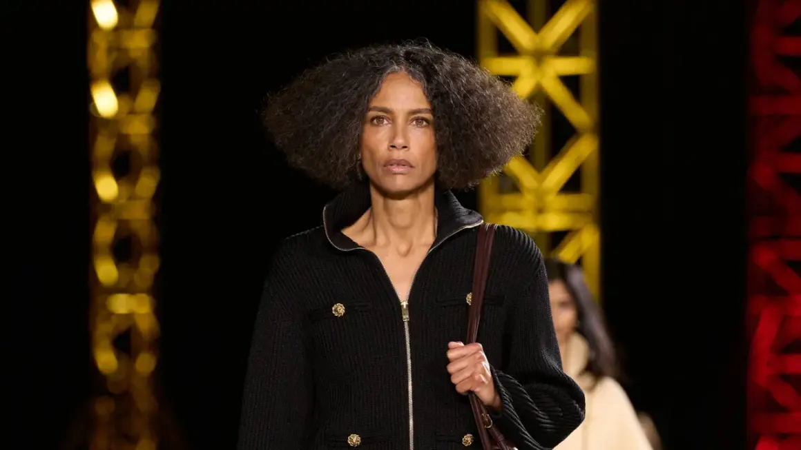 Una mujer con cabello rizado y gris camina por una pasarela, luciendo un elegante cárdigan negro con botones dorados, como una **chanel cantante**.
