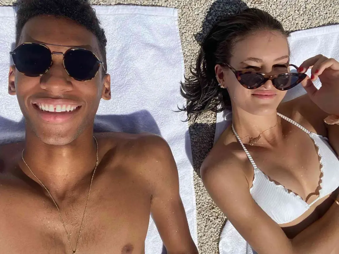 Felix Auger-Aliassime y su novia disfrutan del sol en la playa. Él sonríe a la cámara, ella se ajusta las gafas de sol.