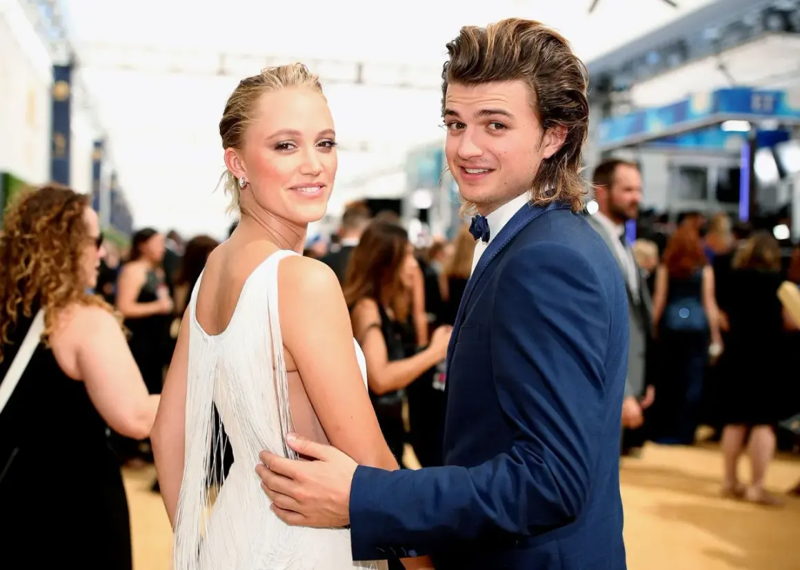 Joe Keery y su pareja, ambos sonriendo a la cámara en una alfombra roja. Ella lleva un vestido blanco con flecos, él un traje azul.