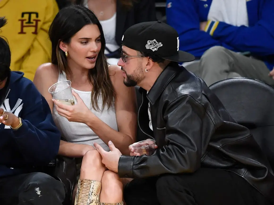 Bad Bunny y Kendall Jenner conversan en un evento deportivo, ella con un vaso en la mano y él con chaqueta de cuero.