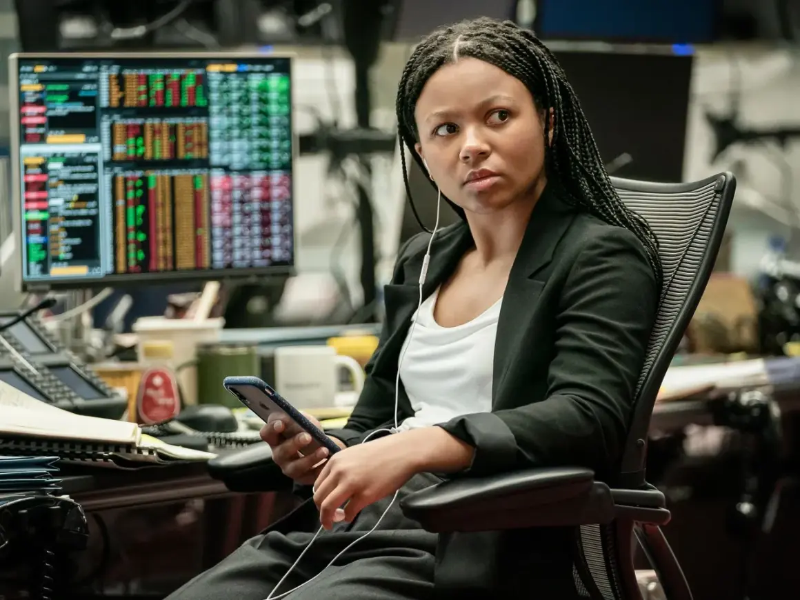 Myha'la Herrold, con auriculares, revisa su teléfono en una oficina con pantallas de datos financieros.