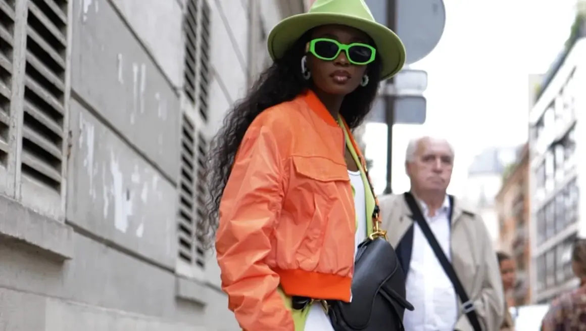 Mujer con sombrero verde y gafas neón, luciendo una bomber naranja vibrante.