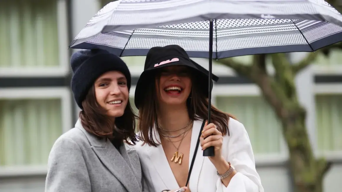 Dos mujeres ríen bajo un paraguas, luciendo un estiloso outfit lluvia mujer con gorros y abrigos.