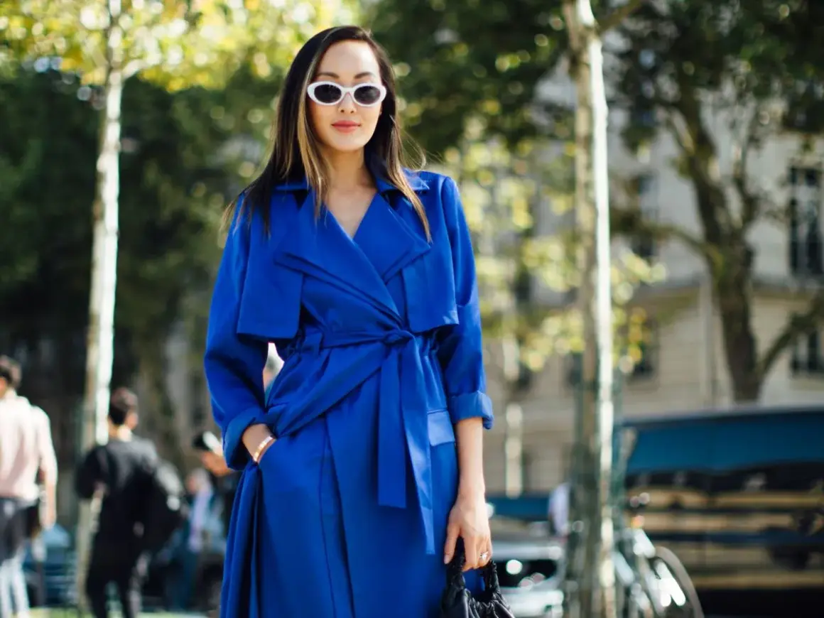 Mujer con gabardina azul eléctrico, gafas blancas y bolso negro, paseando por la calle.