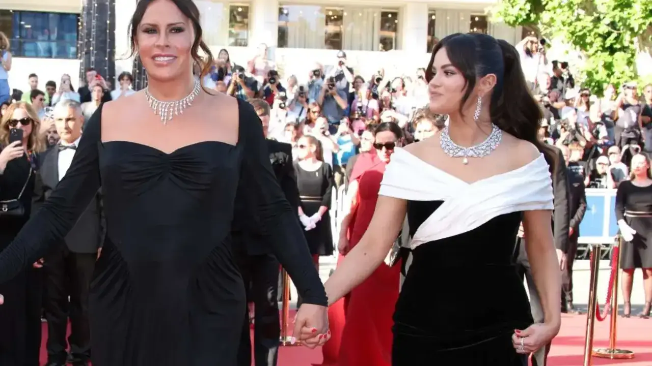 Karla Sofía Gascón y otra mujer, ambas deslumbrantes con vestidos negros y joyas, caminan de la mano por una alfombra roja ante una multitud.