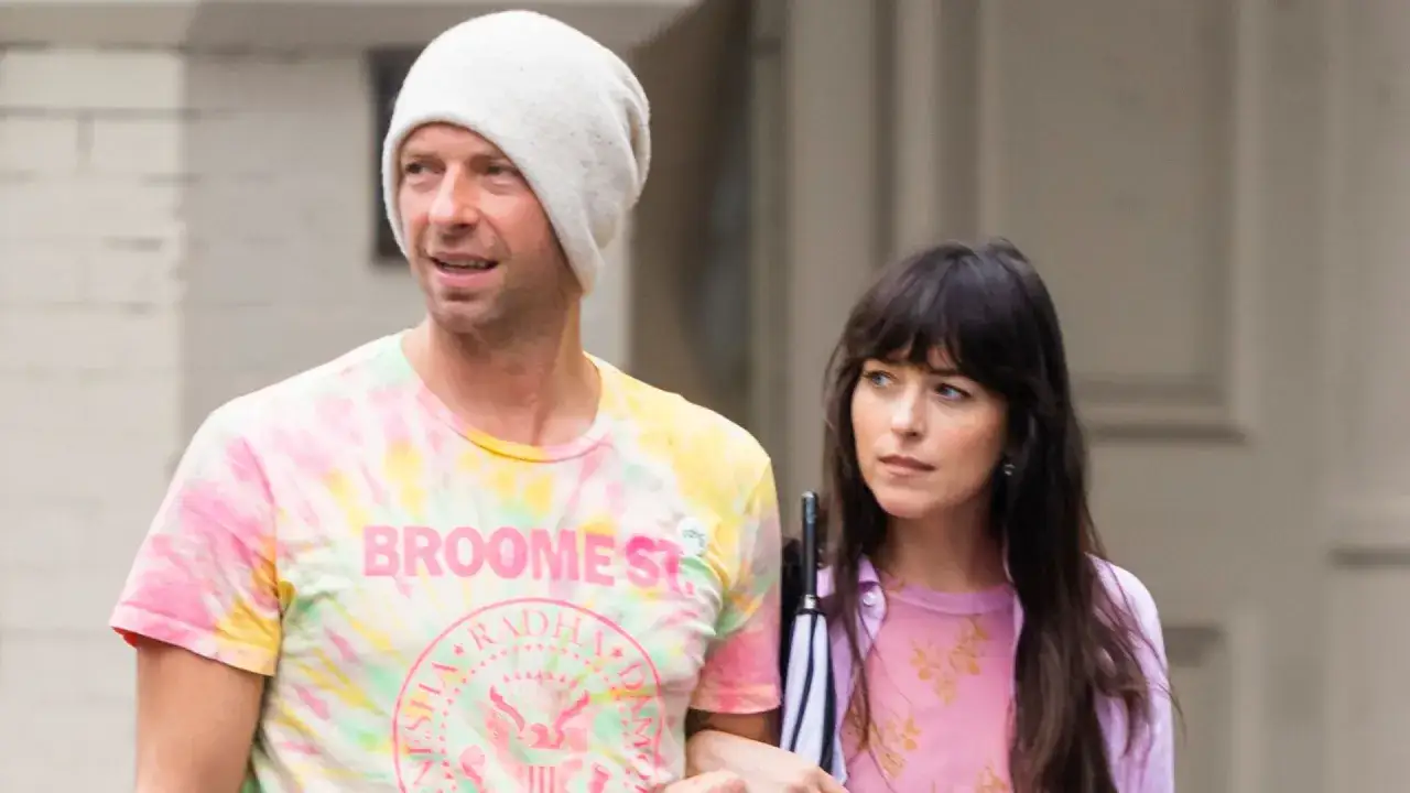 Chris Martin y Dakota Johnson pasean juntos. &Eacute;l lleva una camiseta tie-dye y gorro, ella un vestido rosa.