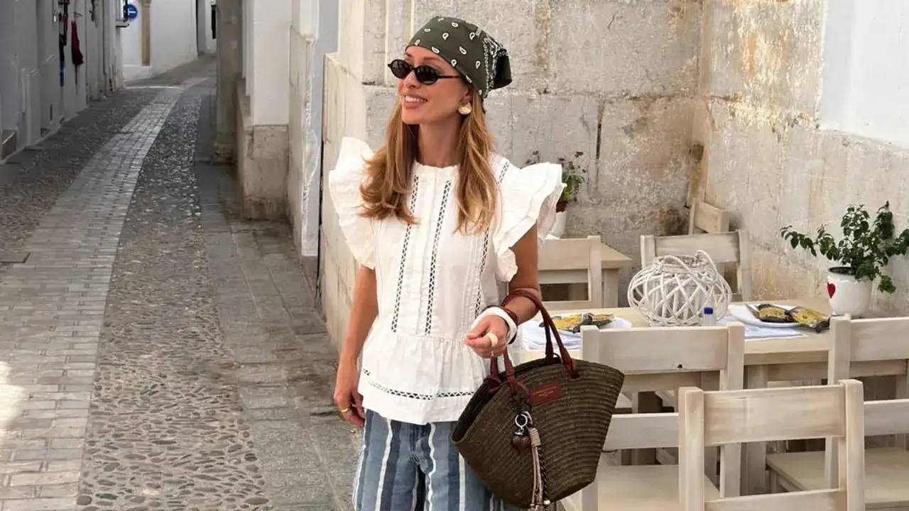 Mujer con pa&ntilde;uelo y gafas de sol, luciendo un top blanco y pantalones a rayas, en un callej&oacute;n empedrado. Su bolso de mimbre complementa su look de street style.