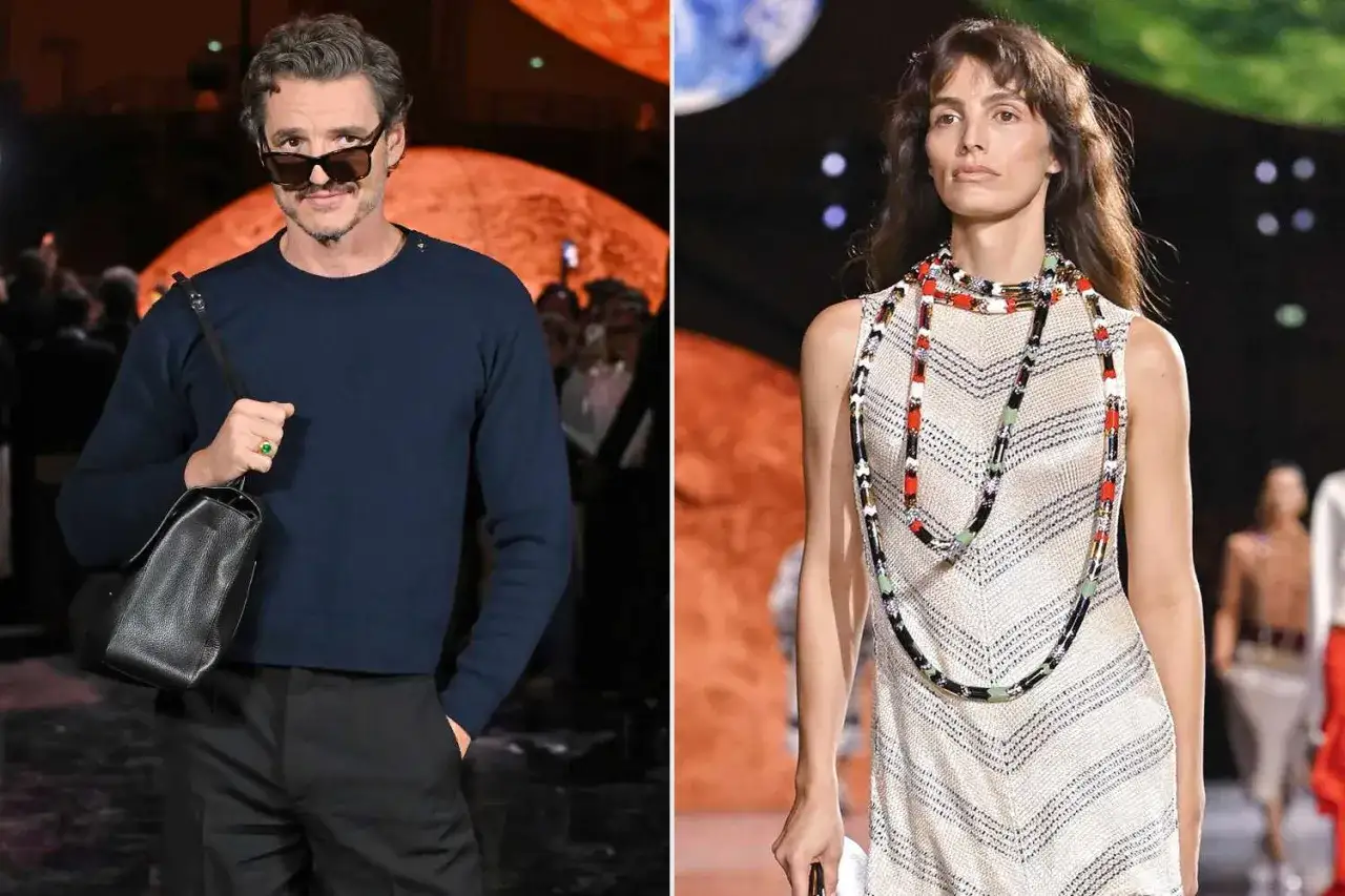 Pedro Pascal, con gafas de sol y un suéter azul, asiste a un evento de moda. A su lado, una modelo luce un vestido blanco con un collar llamativo.