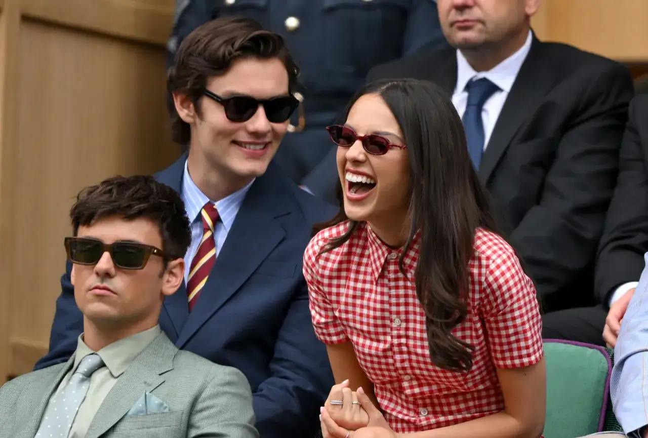 Olivia Rodrigo y su novio, Louis Partridge, disfrutan de un partido en Wimbledon. Ella ríe con entusiasmo, él sonríe.