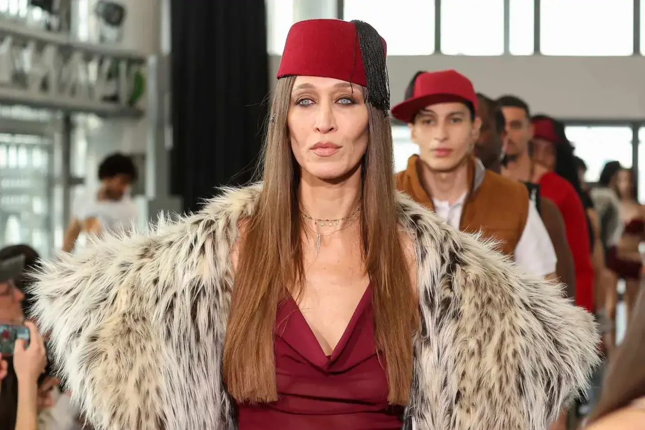 Alana Hadid, con un fez rojo y abrigo de pelo, desfila con otros modelos en una pasarela.
