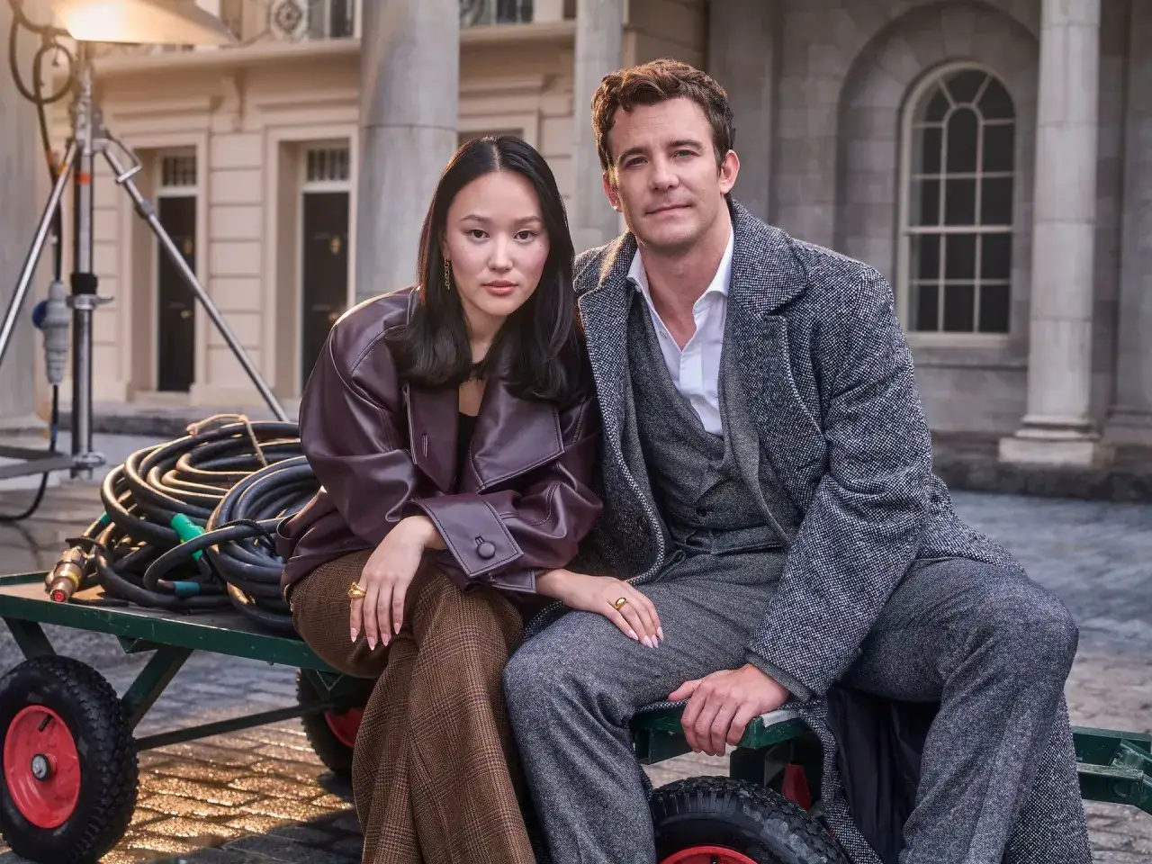 Luke Thompson y su pareja posan sentados en un carrito con cables, con un edificio cl&aacute;sico de fondo.