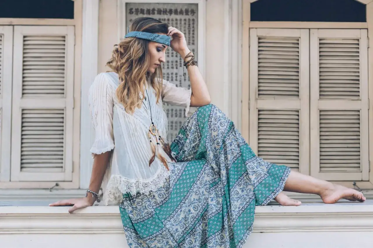 Mujer con estilo boho, diadema azul, blusa blanca y falda larga estampada, sentada en un alf&eacute;izar.