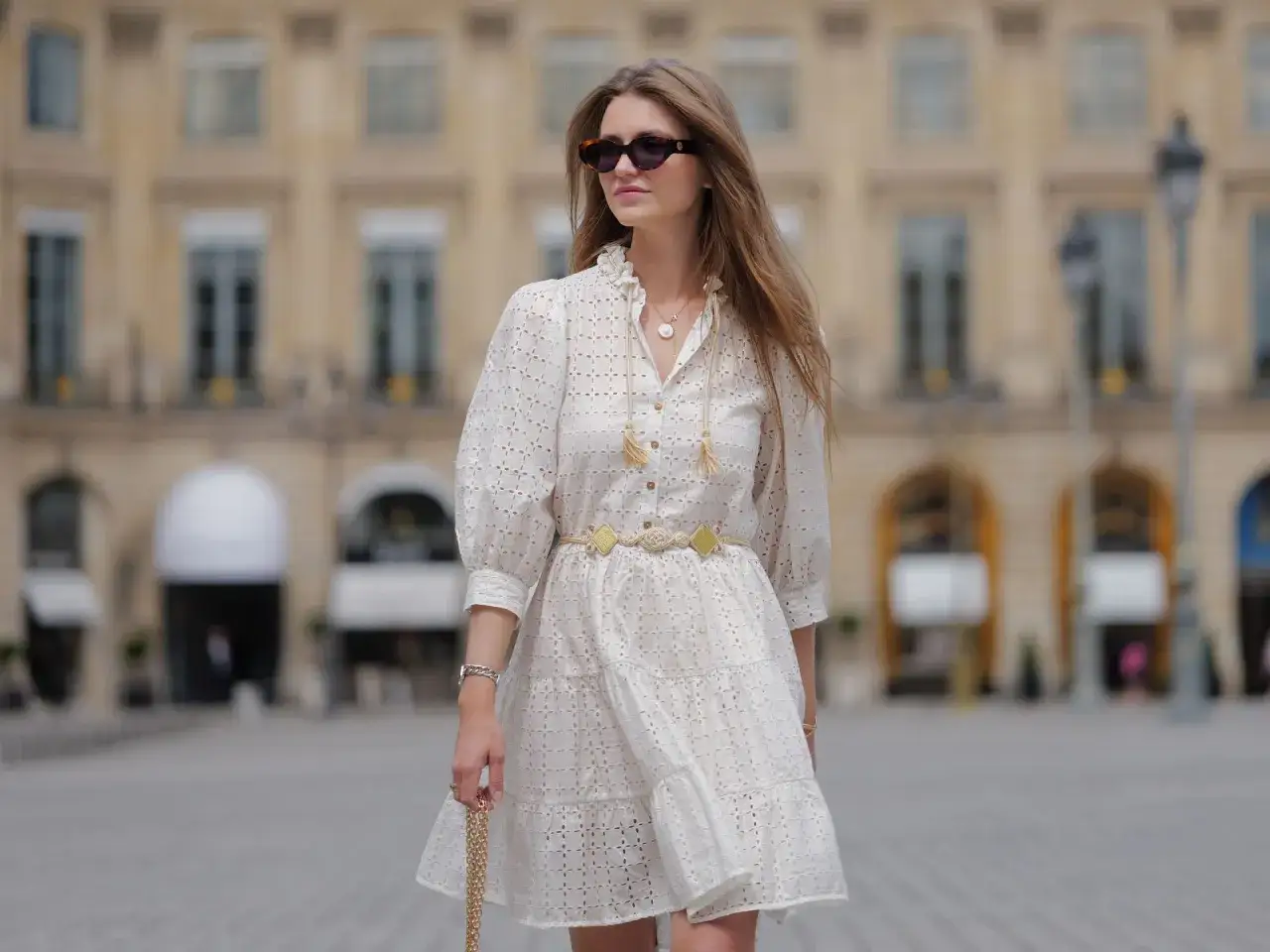 Mujer con vestido blanco calado, cintur&oacute;n dorado y gafas de sol, luciendo un chic estilo franc&eacute;s en Par&iacute;s.