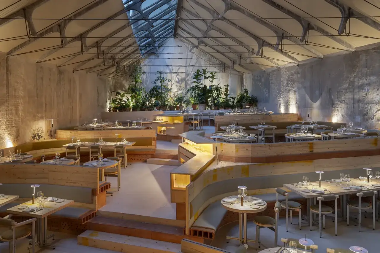 Un restaurante con dise&ntilde;o org&aacute;nico y escalonado, creando un mo de movimiento. Mesas y bancas de madera, plantas y luz tenue.