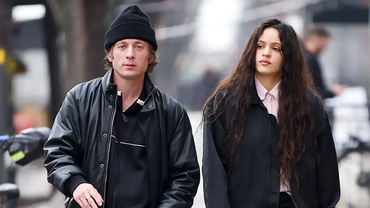 Rosalía y Jeremy Allen caminan juntos, él con gorro y chaqueta de cuero, ella con su larga melena y camisa rosa.