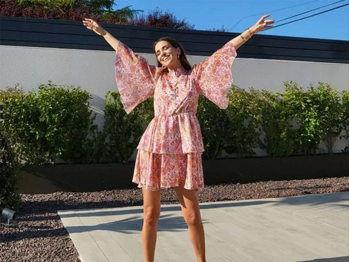 Paula Echevarría, radiante con un vestido rosa estampado, celebra al aire libre con los brazos en alto.