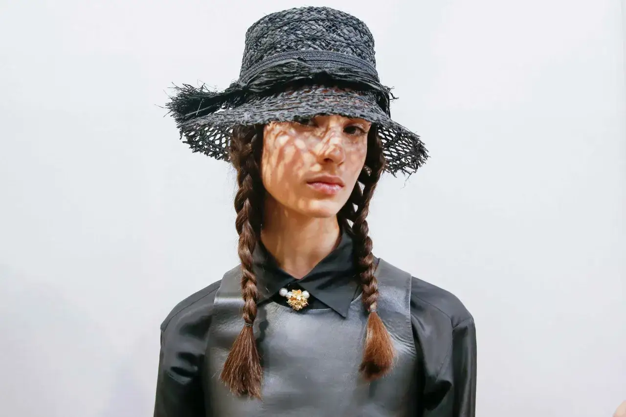 Modelo con sombrero de paja negro y trenzas, luciendo un broche floral, evoca la elegancia de Catherine Dior.