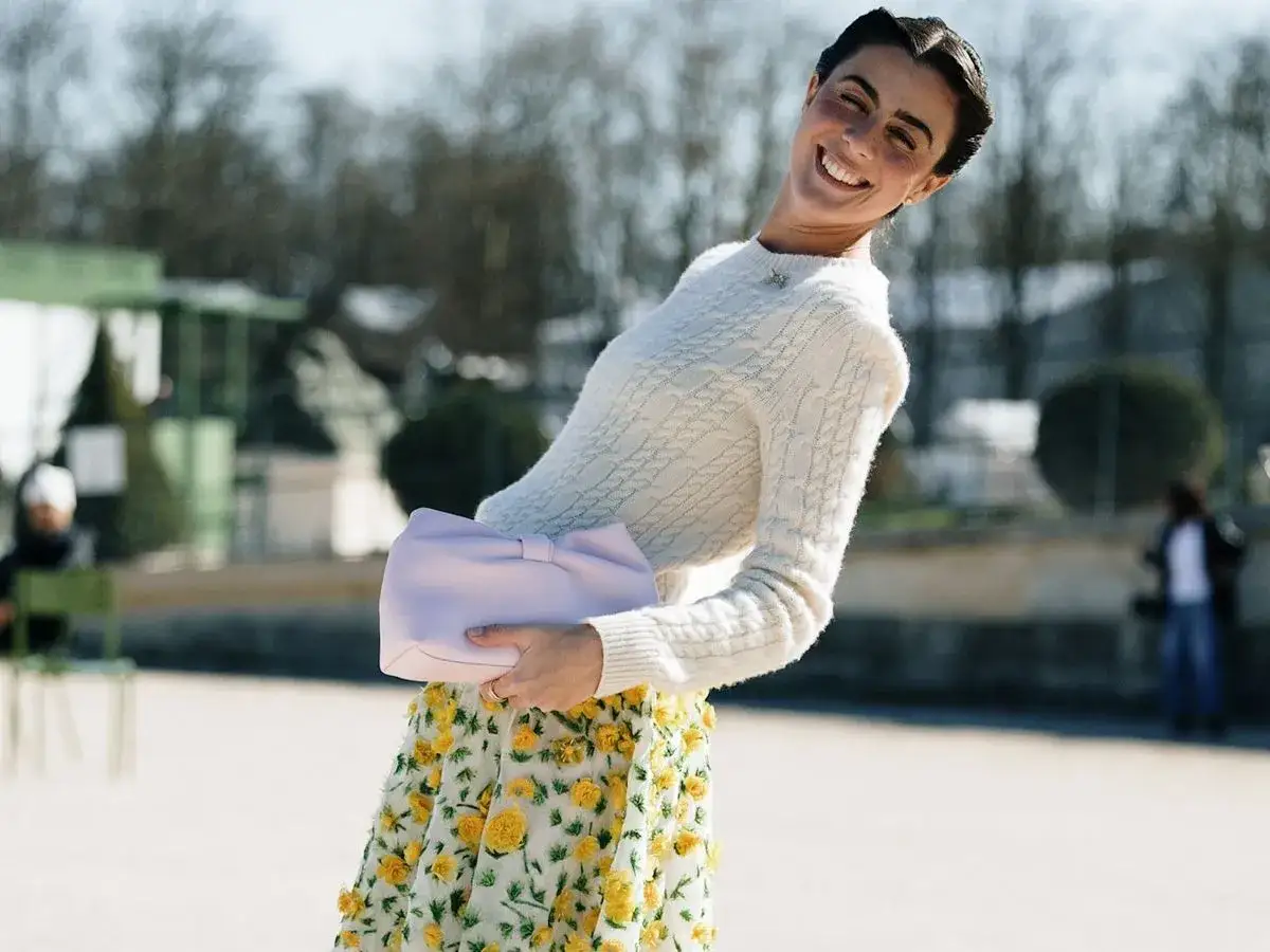 Mujer sonriente con un suéter blanco y falda floral amarilla, perfecta para outfits primavera. Lleva un bolso lila.